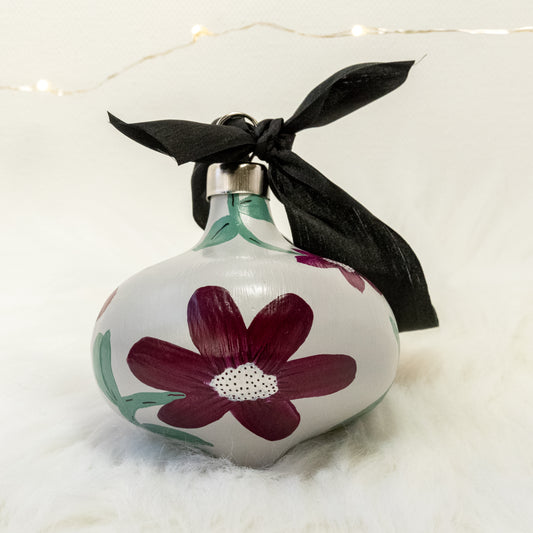The Brenda Hand Painted Ornament features a light gray base coat, deep magenta flowers with sage green stems, white metallic and black accent details. Painted using acryla gouache paints. Displayed on white faux fur with fairy lights in the background.