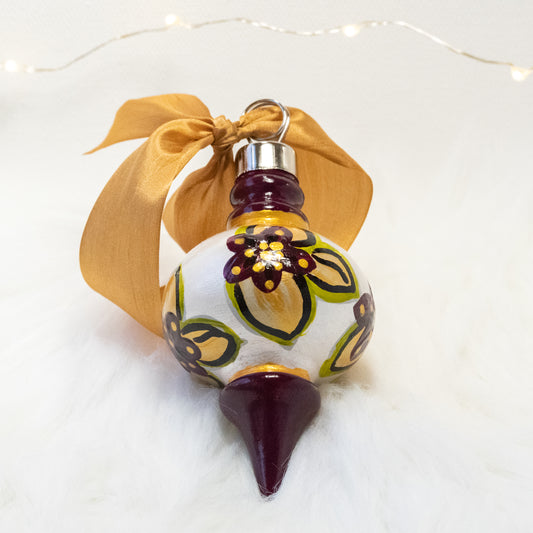 The Britta Hand Painted Ornament features a metallic white and deep magenta base coat, deep magenta flowers with gold and green gold leaves and gold and black accents. Painted using fluid acrylic and acryla gouache paints. Displayed on white faux fur with fairy lights in the background.