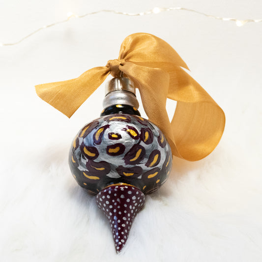 The Brook Lynn Hand Painted Ornament features a black and deep magenta base coat, silver, deep magenta and gold flowers with gold and silver accents. Painted using fluid acrylic and acryla gouache paints. Displayed on white faux fur with fairy lights in the background.