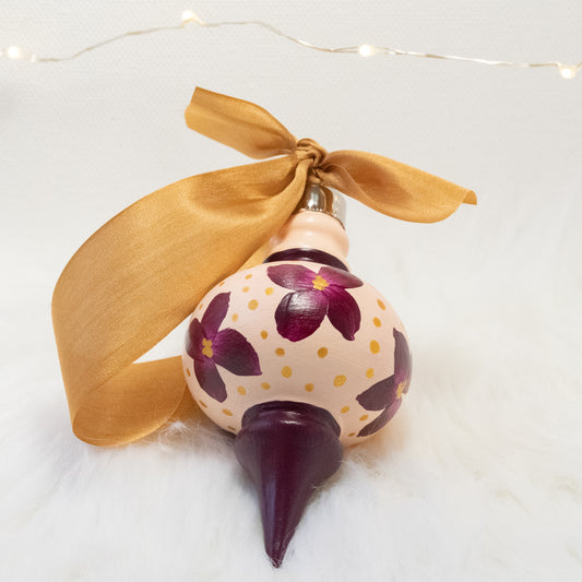The Charlotte Hand Painted Ornament features a light peach and deep magenta base coat, deep magenta flowers with gold polka dots. Painted using fluid acrylic and acryla gouache paints. Displayed on white faux fur with fairy lights in the background.