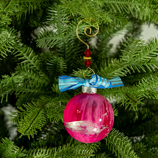 The Melanie Hand Painted Glass Ball Ornament is shades of pink with a hint of white, metallic gold, metallic silver, silver shimmer and a blue striped shimmer ribbon. Painted using fluid acrylic and acrylic paints. Displayed on a Christmas tree.