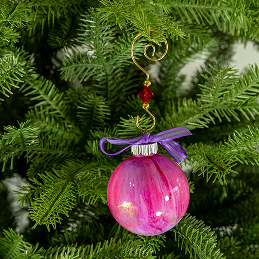 The Katie Hand Painted Glass Ball Ornament has shades of pink and purple with a hint of metallic gold, silver shimmer and a purple satin trimmed ribbon. Painted using fluid acrylic and acrylic paints. Displayed on a Christmas tree.