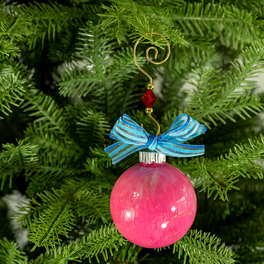 The Sally Hand Painted Glass Ball Ornament is pink with a hint of metallic gold, shimmering silver and a blue striped shimmer ribbon. Painted using fluid acrylic and acrylic paints. Displayed hanging on a Christmas tree.