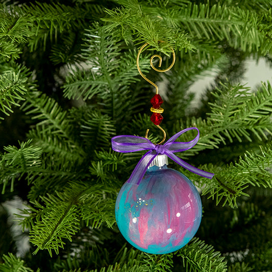 The Erin Hand Painted Glass Ball Ornament is a mix of turquoise blue, lavender and pink with a hint of gold metallic, silver shimmer and a purple satin trimmed ribbon. Painted using fluid acrylic and acrylic paints. Displayed on a Christmas tree.