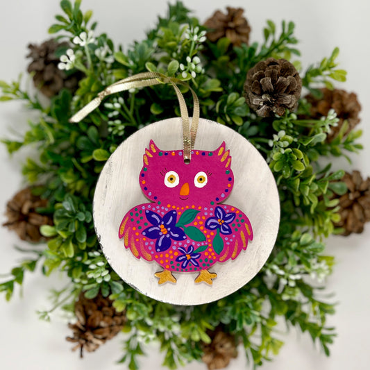 Fuchsia Floral Owl Hand Painted Wood Ornament features a fuchsia pink base with purple & green flowers, orange, green & purple dots with gold feet and feathers displayed on white wood surrounded by greenery and pine cones.