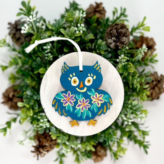 Deep Teal Floral Owl Hand Painted Wood Ornament features a deep teal base with yellow, purple & green flowers and gold feathers & feet displayed on white wood surrounded by greenery and pine cones.
