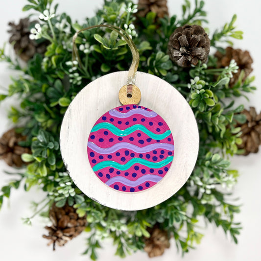 Fuchsia Round Hand Painted Wood Ornament features a fuchsia base with lavender & green wavy stripes, purple dots and a gold cap displayed on white wood surrounded by greenery and pine cones.