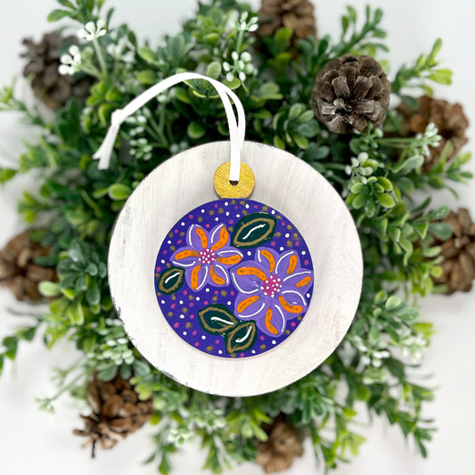Deep Purple Floral Hand Painted Round Wood Ornament features a deep purple base with lavender, orange, pink & white flowers with green leaves and pink, gold & white dots with a gold cap displayed on white wood surrounded by greenery and pine cones.