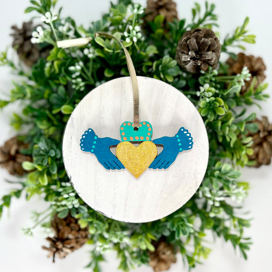 Deep Teal Irish Claddagh Hand Painted Wood Ornament features deep teal hands with green & white dots on the cuffs, an aqua green crown with orange dots and a gold heart displayed on white wood surrounded by greenery and pine cones.