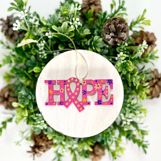 Fuchsia, Purple and Orange Hope Ribbon Hand Painted Wood Ornament features a fuchsia background with orange & purple flowers and dots and gold dots inside the ribbon displayed on white wood surrounded by greenery and pine cones.