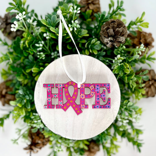 Fuchsia, White, Yellow and Purple Hope Ribbon Hand Painted Wood Ornament features a fuchsia background with white, yellow and purple flowers along with aqua, purple and blue dots and gold dots inside the ribbon displayed on white wood surrounded by greenery and pine cones.