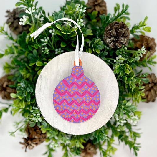 Purple, Pink & Orange Round Hand Painted Wood Ornament features purple with pink zig zags, orange polka dots and an orange cap displayed on white wood surrounded by greenery and pine cones.