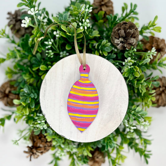 Purple Colorful Striped Bulb Hand Painted Wood Ornament features purple with yellow, pink & orange stripes displayed on white wood surrounded by greenery and pine cones.