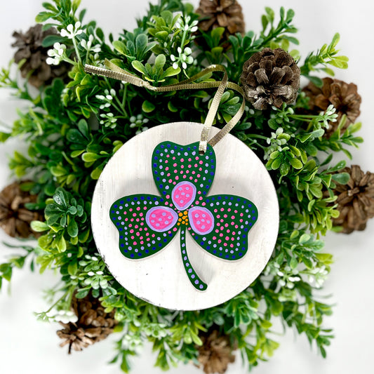 The Dark Green Shamrock Hand Painted Wood Ornament features a dark green base with a pink & purple flower, as well as pink & purple dots displayed on white wood surrounded by greenery and pine cones.