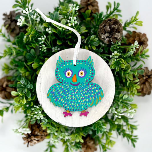 Aqua Owl Hand Painted Wood Ornament features an aqua base with yellow, purple & gold feathers, orange with fuchsia feet displayed on white wood surrounded by greenery and pine cones.