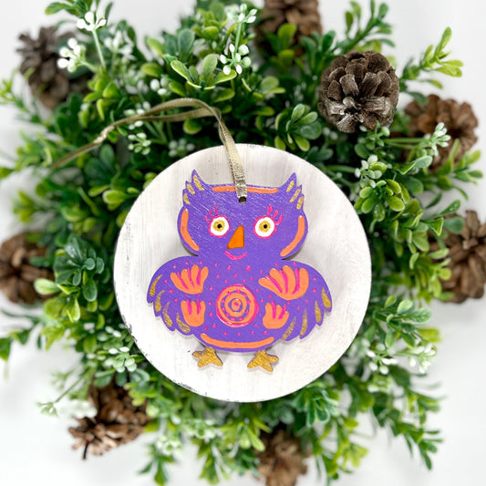 Purple Floral Owl Hand Painted Wood Ornament features a purple base with orange & pink flowers and pink dots and gold feathers & feet displayed on white wood surrounded by greenery and pine cones.