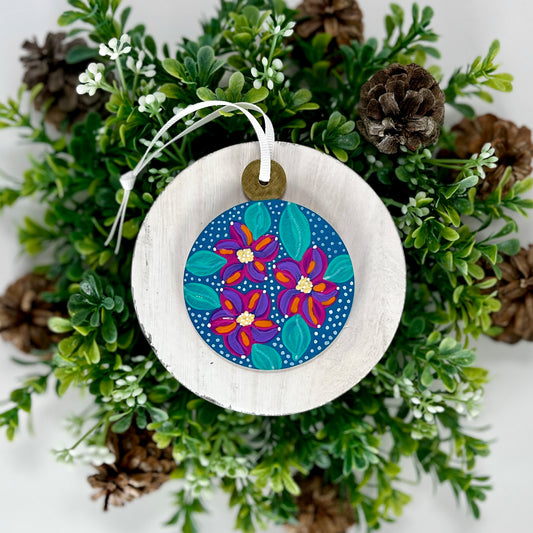 Blue Floral Hand Painted Round Wood Ornament features a blue base with lavender, pink & orange flowers with aqua green leaves, white polka dots and a gold cap displayed on white wood surrounded by greenery and pine cones.
