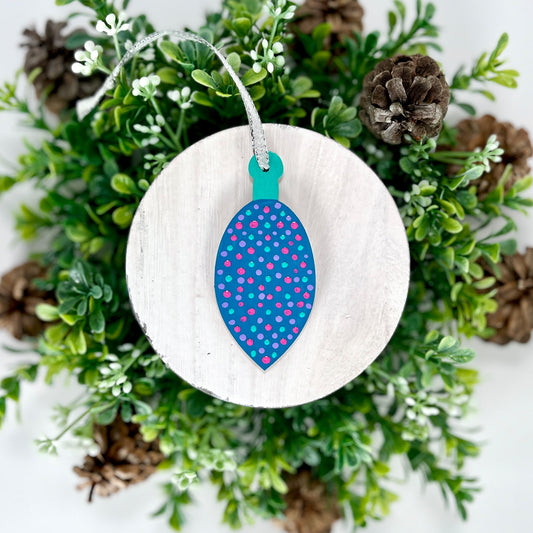 Blue Polka Dot Hand Painted Bulb Wood Ornament features a blue base with lavender, pink & aqua green polka dots and an aqua green cap displayed on white wood surrounded by greenery and pine cones.