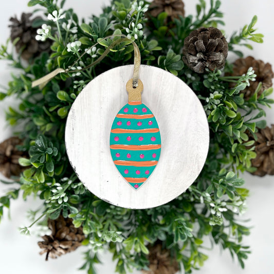 Aqua Green Striped Polka Dot Hand Painted Bulb Wood Ornament features an aqua green base with orange stripes and pink polka dots and a gold cap displayed on white wood surrounded by greenery and pine cones.