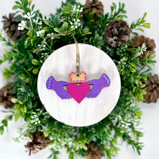 Purple Irish Claddagh Hand Painted Wood Ornament features purple hands with pink & white dots on the cuffs, a gold crown with pink dots and a pink heart displayed on white wood surrounded by greenery and pine cones.