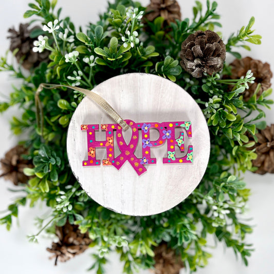 Fuchsia, Orange, Purple and Green Hope Ribbon Hand Painted Wood Ornament features a fuchsia background with orange, purple and green flowers along with aqua, yellow and orange dots and gold dots inside the ribbon displayed on white wood surrounded by greenery and pine cones.
