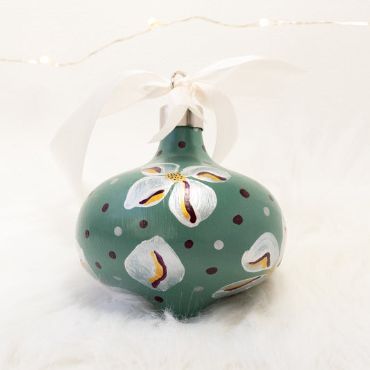 The Kristina Hand Painted Ornament features a sage green base coat, metallic white flowers with deep magenta and gold accent details. Painted using acryla gouache paints. Displayed on white faux fur with fairy lights in the background.